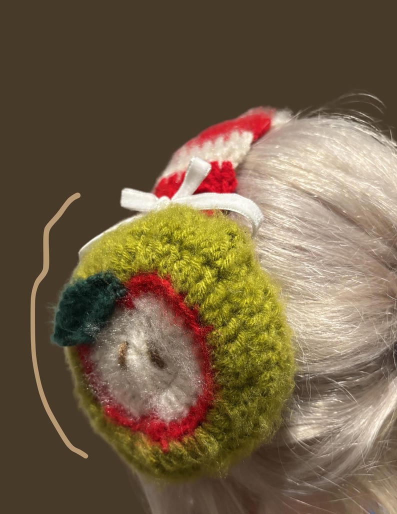 May include: A handmade, crocheted apple-shaped hair accessory. The apple is primarily green with a red outline, white center, and a small green leaf. A red and white striped band and white ribbon complete the design.