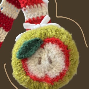 May include: A handmade crochet apple ornament with a green exterior, red interior, and a white core. The apple has a green leaf and is attached to a red and white striped stem. A white ribbon is tied near the apple.