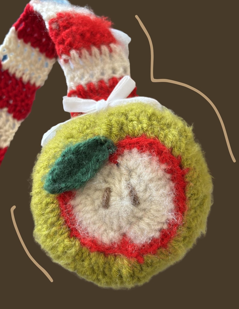 May include: A handmade crochet apple ornament with a green exterior, red interior, and a white core. The apple has a green leaf and is attached to a red and white striped stem. A white ribbon is tied near the apple.