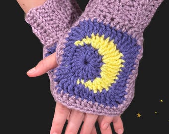 May include: Crocheted fingerless gloves in lavender with a square design featuring a blue circle and a yellow crescent moon. The gloves are handmade and appear soft and warm, perfect for cooler weather.