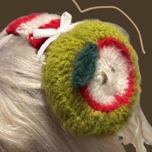 May include: A handmade green apple-shaped earmuff with a red and white striped top and a white bow. The apple design features a red outline, white center, and a green leaf. The earmuffs are designed to be worn on the head.