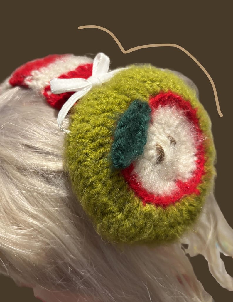 May include: A handmade green apple-shaped earmuff with a red and white striped top and a white bow. The apple design features a red outline, white center, and a green leaf. The earmuffs are designed to be worn on the head.