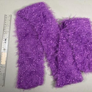 May include: A pair of vibrant purple leg warmers made of a fuzzy, textured material. The leg warmers are a solid color with a speckled pattern and are lying flat on a light-colored surface. A ruler is placed next to the leg warmers.