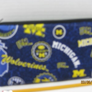 May include: A rectangular pencil case with a black zipper. The case is navy blue with a pattern of yellow and white Michigan Wolverines logos and text. The pencil case is approximately 8 inches long.