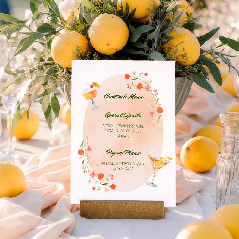 Op de afbeelding: Een cocktailmenukaart met de tekst "Cocktail Menu" en twee cocktailillustraties. Het menu bevat Aperol Spritz en Paper Plane cocktails, met ingredi&euml;nten. De kaart staat op een houten standaard, omringd door citroenen en groen.