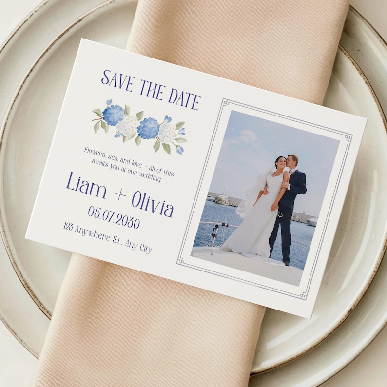 Puede incluir: Una tarjeta blanca de 'Save the Date' con dise&ntilde;os florales azules y una foto de una pareja. La tarjeta muestra los nombres Liam y Olivia, la fecha 05.07.2030 y una direcci&oacute;n. Descansa sobre una servilleta beige y un plato blanco.