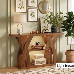 May include: A light brown wooden console table with an X-shaped base and a middle shelf. The table holds a lamp with a beige shade, three small round decorative objects, a stack of books, a camera, and two white rectangular storage boxes.