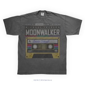 May include: Dark gray t-shirt featuring a cassette tape design with the text "MOONWALKER" and "MICHAEL JACKSON". The design includes the text "Smooth Criminal" and the years 1958 and 2009. The shirt is a crew neck.