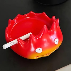 Pode incluir: Um cinzeiro vermelho e amarelo em forma de demónio de fogo de desenho animado com olhos brancos. Um cigarro branco repousa no cinzeiro. O cinzeiro está sobre uma superfície preta.