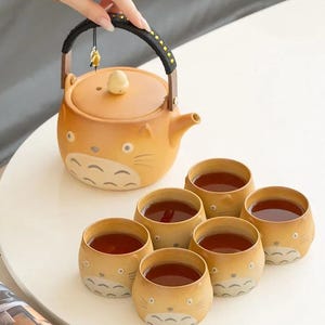 Puede incluir: Juego de té de cerámica de color naranja claro que incluye una tetera y seis tazas a juego, cada una con caras de estilo caricatura. La tetera tiene un asa de madera con un agarre negro. Las tazas están llenas de un líquido oscuro.