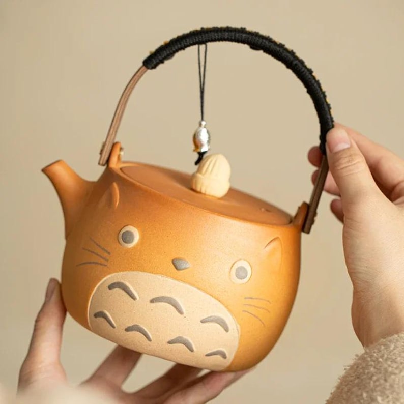May include: A ceramic teapot in the shape of a cartoon character, featuring a brown body with gray and beige accents. The teapot has a black handle and a small charm hanging from the lid. The teapot is being held by a person.
