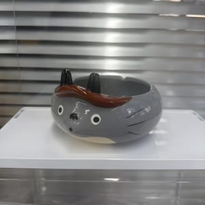 以下が含まれることがあります： 黒い耳と額の茶色のストライプが付いた、漫画のキャラクターの形をした灰色のセラミック灰皿。 灰皿は、円形の開口部と2つの小さな白い円の目を持つ丸いボディをしています。