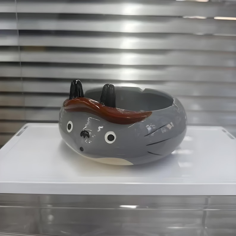 以下が含まれることがあります： 黒い耳と額の茶色のストライプが付いた、漫画のキャラクターの形をした灰色のセラミック灰皿。 灰皿は、円形の開口部と2つの小さな白い円の目を持つ丸いボディをしています。