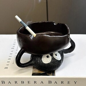 Pode incluir: Um cinzeiro de cer&acirc;mica castanho escuro com um cigarro, pousado sobre uma estatueta preta com olhos brancos. O cinzeiro tem uma forma curva com uma pega de cada lado. A fuma&ccedil;a sobe do cigarro. A estatueta est&aacute; pousada num livro.