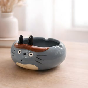 Puede incluir: Cenicero de cerámica gris con forma de personaje de dibujos animados, con orejas negras y una banda marrón. El cenicero tiene forma redonda con un área empotrada para sostener cenizas y cigarrillos. El personaje tiene ojos blancos y bigotes negros.