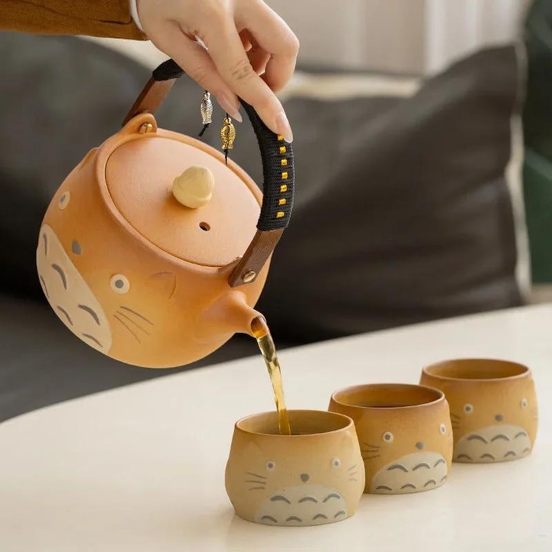 May include: A ceramic teapot and three matching cups are depicted. The teapot, with a brown handle and lid, is pouring tea into a cup. The teapot and cups feature a cartoon cat design in shades of brown and beige. The tea set is on a white surface.