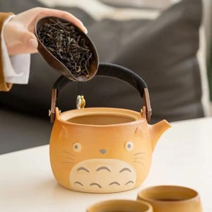 May include: A ceramic teapot with a cartoon cat design in shades of orange and cream. The teapot has a black handle and a small spout. A wooden scoop pours tea leaves into the teapot. Two small teacups are in the foreground.