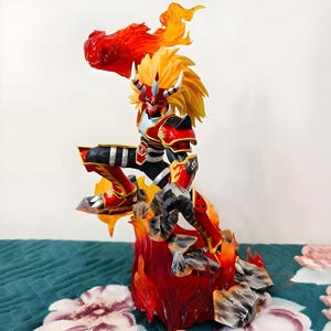 Peut inclure: Figurine détaillée d'un personnage en pose dynamique, avec une armure rouge et noire avec des accents dorés. La figurine a des cheveux jaunes et un effet de feu émanant de la tête et de la base. La base comprend des structures rocheuses.