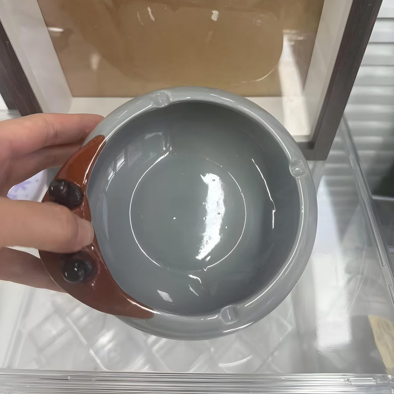 以下が含まれることがあります： 丸い灰皿。グレーのセラミック製で、茶色の三日月型のアクセントが付いています。灰皿には、タバコを置くための3つの小さな溝があります。シンプルでモダンなデザインで、家庭やオフィスでの使用に適しています。
