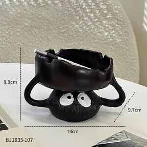 Pode incluir: Cinzeiro preto com design de desenho animado, apresentando uma base redonda com olhos brancos e um topo em forma de tigela marrom escuro. Mede 14 cm de largura, 9,7 cm de altura e 8,8 cm de profundidade. Um cigarro repousa no topo. C&oacute;digo do produto: BJ1835-107.