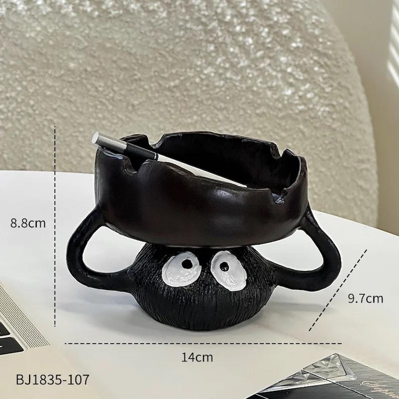 Pode incluir: Cinzeiro preto com design de desenho animado, apresentando uma base redonda com olhos brancos e um topo em forma de tigela marrom escuro. Mede 14 cm de largura, 9,7 cm de altura e 8,8 cm de profundidade. Um cigarro repousa no topo. C&oacute;digo do produto: BJ1835-107.