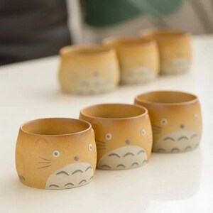 May include: A set of six ceramic cups with a cartoon cat design. The cups are cylindrical with a light brown exterior and a lighter interior. The cat face features gray accents and is centered on the cup's front.