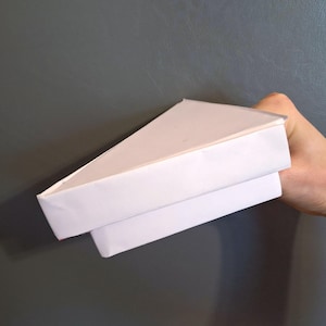 Puede incluir: Un triángulo de origami de papel blanco sostenido sobre un fondo gris. El origami está doblado en forma triangular, con una segunda capa de papel debajo. Los bordes son limpios y nítidos.