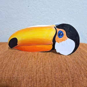 May include: A colorful toucan head sculpture with a large yellow beak, black and white head, and blue eye. The beak has a black spot and orange underside. The sculpture is resting on a brown surface.