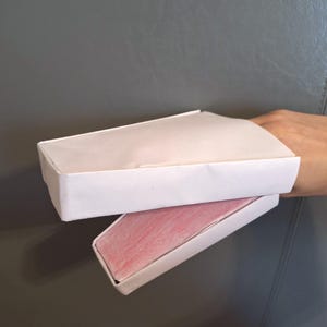 Puede incluir: Dos cajas rectangulares blancas de papel, una encima de la otra, sostenidas por una mano. La caja superior está cerrada, mientras que la caja inferior revela un interior rosa. Las cajas son de papel.