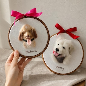 May include: Two circular wooden embroidery hoops containing needle-felted dog portraits on white fabric. One features a tan dog named Madonna with a pink bow, the other a white dog named Harry with a red bow. Both dogs have tongues sticking out.