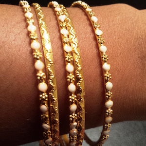 Puede incluir: Se muestran cuatro pulseras doradas con detalles blancos. Las pulseras presentan un diseño texturizado y están adornadas con pequeñas cuentas blancas. Las joyas se llevan en un brazo, destacando el intrincado diseño de las pulseras.