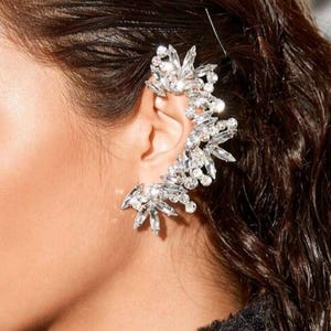 Puede incluir: Un brazalete de oreja brillante con un diseño floral de cristales transparentes. La joya se lleva en la oreja y se coloca sobre cabello castaño oscuro. Los cristales reflejan la luz, creando un aspecto glamuroso.