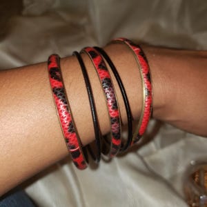 May include: A collection of bracelets in the image. The bracelets include several black and red bangles with a snakeskin pattern, and several thin black bracelets. The bracelets are stacked on a wrist.