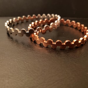 Könnte beinhalten: Zwei Metallarmreifen mit einem wellenförmigen Design. Einer ist silberfarben und der andere kupferfarben. Die Armreifen liegen auf einer dunklen, strukturierten Oberfläche.