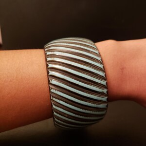 May include: A wide, striped bangle bracelet. The bracelet features alternating bands of light blue and dark brown. The bracelet is worn on a wrist and has a textured, ridged surface.