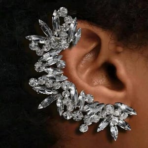 May include: A sparkling ear cuff featuring a floral design. The cuff is adorned with numerous clear, faceted crystals of varying sizes, creating a dazzling, eye-catching accessory. The crystals are arranged to mimic the shape of leaves and flowers.