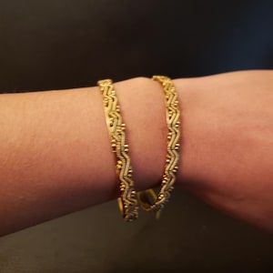 Op de afbeelding: Goudkleurige armband met een ingewikkeld geweven ontwerp. De armband heeft een reeks kleine, ronde kralen en een gedetailleerd patroon. De sieraden worden op de arm van een persoon getoond, waarbij het ontwerp en de textuur van de armband worden getoond.