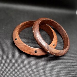 May include: Two wooden bangles with a rich brown color. Each bangle is adorned with small, dark metal studs. The bangles are circular and have a smooth, polished finish, showcasing the natural wood grain.
