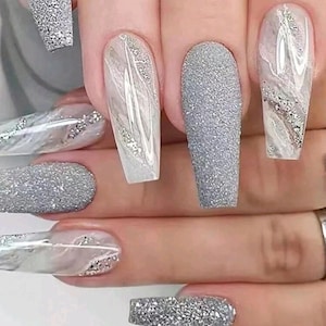May include: Close-up of a hand with long, coffin-shaped acrylic nails. The nails feature a variety of designs, including glitter, marble effects, and silver accents. The color palette is primarily silver and white, creating a glamorous look.