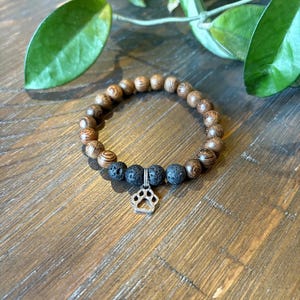 May include: A bracelet made of wooden beads, lava beads, and a silver paw print charm. The bracelet is displayed on a wooden surface. The beads are brown and black. Green leaves are in the background.
