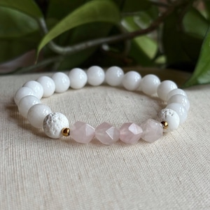 May include: A white beaded bracelet with a section of faceted pink beads and gold-coloured accents. The bracelet features a mix of smooth and textured white beads. The background includes green foliage.