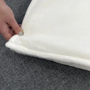 May include: A close-up of a white, plush blanket with a soft, textured surface. The blanket has a thick, stitched border. The blanket is being held by a hand with a gray nail polish.
