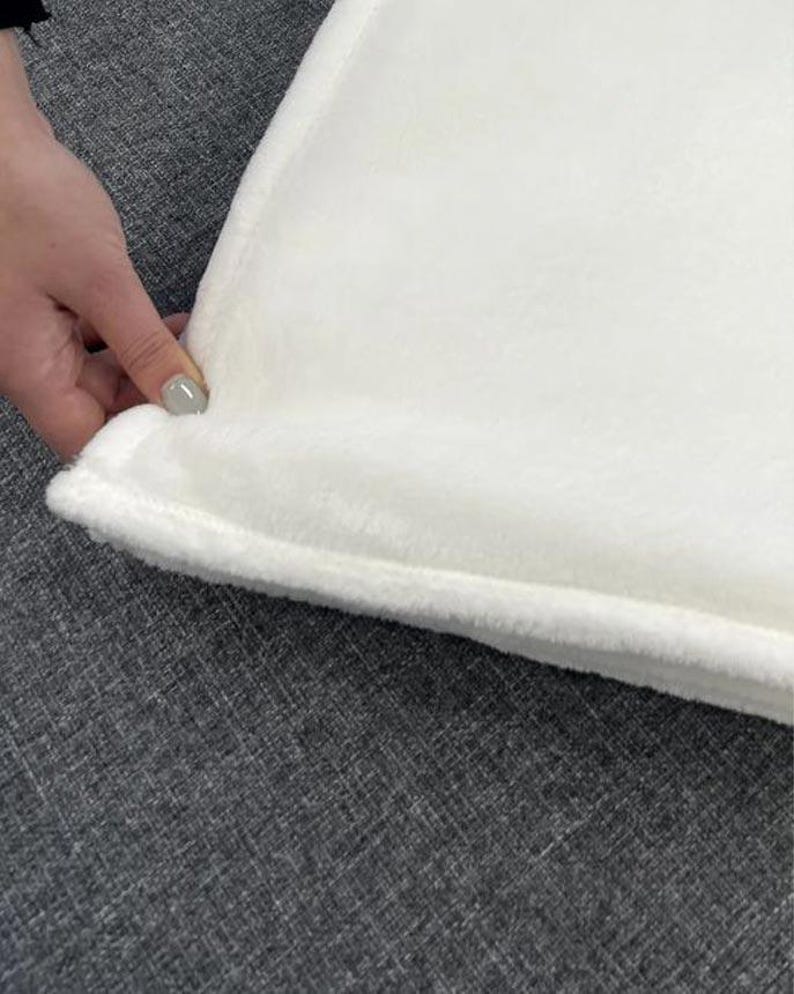 May include: A close-up of a white, plush blanket with a soft, textured surface. The blanket has a thick, stitched border. The blanket is being held by a hand with a gray nail polish.