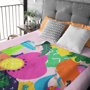 May include: A colorful blanket with a vibrant fruit and floral design. The blanket features large pink, orange, yellow, and blue shapes, including a lemon slice and a stylized figure, set against a light pink background.
