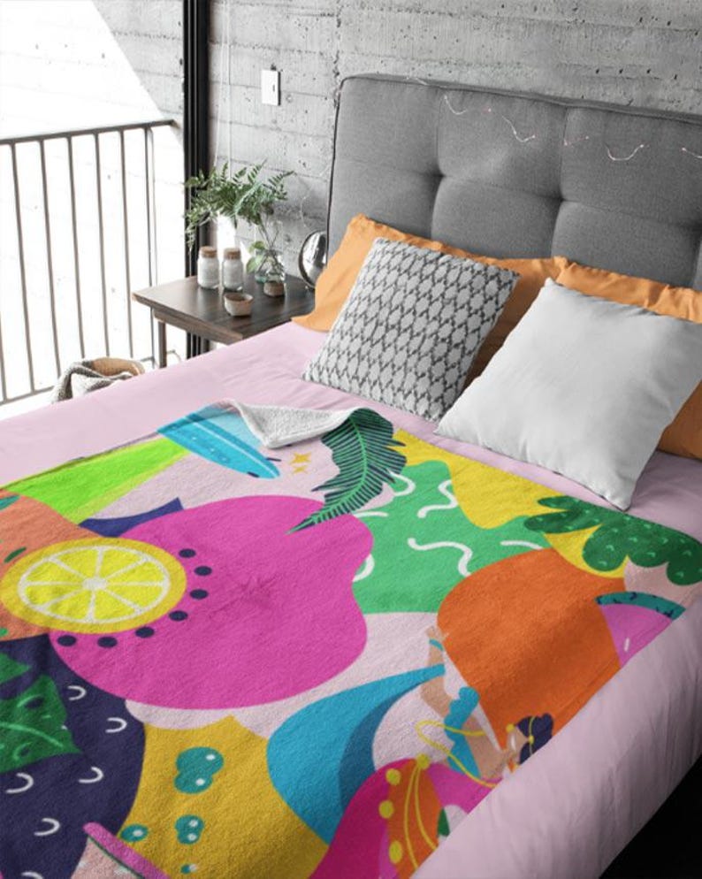 May include: A colorful blanket with a vibrant fruit and floral design. The blanket features large pink, orange, yellow, and blue shapes, including a lemon slice and a stylized figure, set against a light pink background.