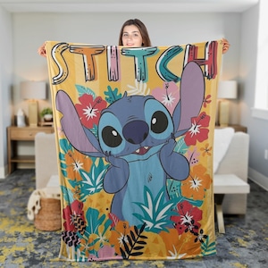 May include: A yellow blanket featuring the word "STITCH" in colorful letters. The blanket showcases a cartoon image of Stitch surrounded by tropical flowers in red, pink, and orange. The blanket is held up by a person.