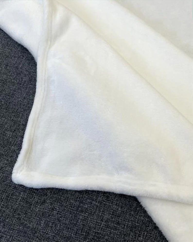 May include: A close-up shot of a soft, white blanket. The blanket is folded, revealing a plush texture and a smooth, finished edge. The fabric appears to be a cozy, warm material, perfect for snuggling. The blanket is a solid, bright white color.