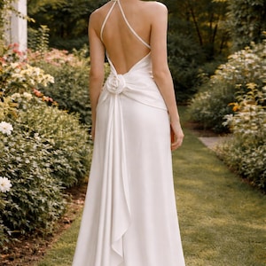 May include: A white halter-neck wedding gown with a low back and a floral embellishment at the waist. The dress features a flowing skirt and a train. The model is standing in a garden setting.