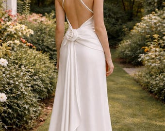 May include: A white halter-neck wedding gown with a low back and a floral embellishment at the waist. The dress features a flowing skirt and a train. The model is standing in a garden setting.