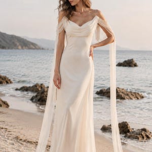 May include: A floor-length ivory gown featuring a draped cowl neckline, thin spaghetti straps, and sheer off-the-shoulder sleeves that extend into long, flowing fabric panels. The dress has a fitted bodice and a smooth, floor-length skirt.